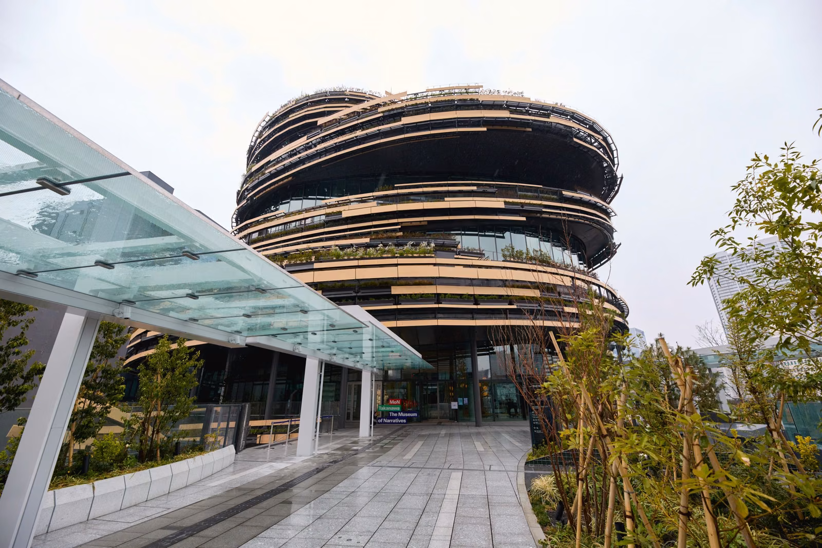 MoN Takanawa: The Museum of Narratives の外観。木の水平ルーバーと植栽が幾重にも巡る円形の建物と、ガラス屋根の続く入口